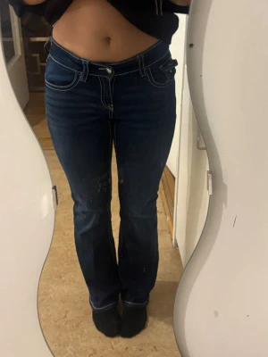 Mörkblå bootcut jeans med kontrastsömmar - Säljer ett par mörkblå jeans med tydliga vita kontrastsömmar och bootcut-modell. Byxorna har klassiska fem fickor, knappstängning och dekorativ knapp på bakfickan. Snygg passform som markerar midjan och lätt utsvängda ben.
