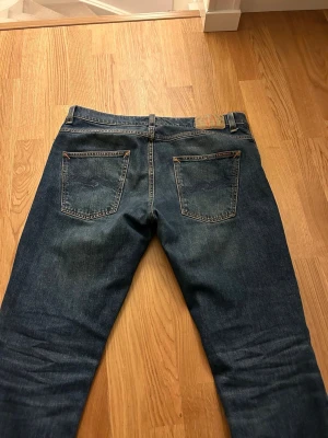Blå jeans från Nudie Jeans, W33 L30 - Säljer ett par klassiska blå jeans från Nudie Jeans med snygga detaljer på bakfickorna och orange sömmar. Modellen har rak passform och normal midja, tillverkade i slitstarkt bomullsmaterial. Perfekt för dig som gillar tidlös stil och vill ha jeans som funkar till allt.