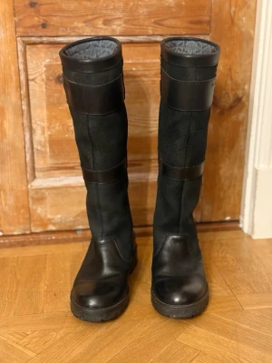 Svarta Dubarry Gore-Tex stövlar - Dubarry of Ireland svarta stövlar med Gore-Tex-foder, tillverkade i slitstarkt skinn och mocka. Stövlarna har två snygga spännen upptill, grov platt sula och rund tå. Insidan är fodrad med grått mönstrat tyg för extra komfort. Perfekta för dig som gillar stil och funktion. Nypris ca 4700kr