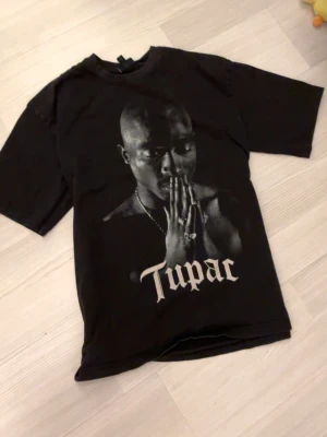 Svart 2Pac t-shirt med tryck - Svart t-shirt med stort tryck av Tupac på framsidan och texten 'Tupac' i vit stil. Klassisk rund hals och korta ärmar. Perfekt för dig som gillar streetwear och vill ha en ikonisk look.