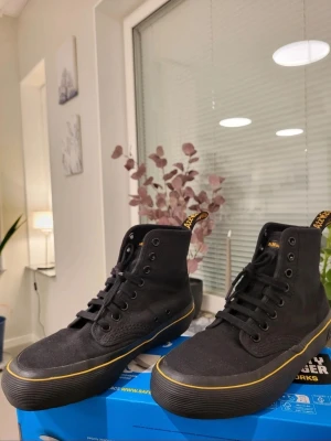 Svarta Dr. Martens Monet sneakers 36 - Säljer ett par svarta höga sneakers från Dr Martens med snygga gula detaljer längs sulan och på hälen. Skorna har snörning, canvasmaterial och en platt sula. Perfekta för dig som gillar streetstyle och vill ha något som sticker ut lite extra.
