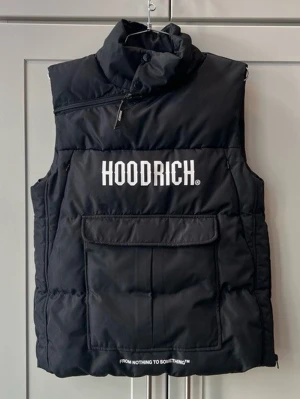 Svart dunväst från Hoodrich - Säljer en svart dunväst från Hoodrich med hög krage och stor ficka framtill. Västen har vit logga på bröstet och textdetalj längst ner. Perfekt för lager på lager och streetwear-stil.