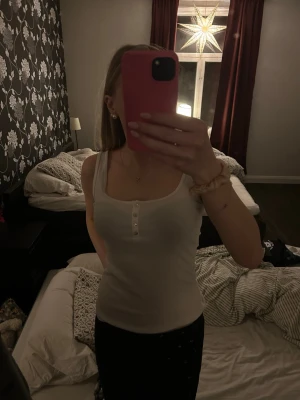 Vit ribbad linnetopp från H&M - Säljer en enkel och snygg vit ribbad linnetopp från H&M. Toppen har smala axelband, rundad halsringning och dekorativa pärlemorknappar framtill. Perfekt att styla med jeans eller kjol för en clean look.