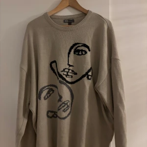 Beige stickad tröja med ansiktsmotiv - Säljer en oversized beige stickad tröja från ASOS med ett svart grafiskt ansiktsmotiv på framsidan. Tröjan har rund halsringning och långa ärmar, perfekt för lager-på-lager under kyliga dagar.