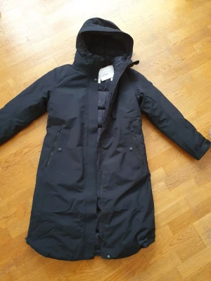 Svart lång dunparkas med huva - Säljer en svart lång parkas från Ahkka  med huva (avtagbar pälskant). Dragkedjor på sidorna för ökad rörlighet samt tvåvägs dragkedja framtill. Jackan är varm och go', går att dra åt i midjan. Vattenavvisande. En toppenjacka men jag ångrade mig (för sent att skicka tbx vid det laget). Perfekt för kalla vinterdagar. Oanvänd med lappar kvar.  Nypris 2900, säljer den för 1900kr