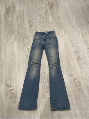 Blå bootcut jeans med slitningar - Säljer ett par ljusblå bootcut jeans från Nelly med slitna detaljer över knäna och snygg tvätt. Jeansen har klassiska fickor både fram och bak, samt knapp och dragkedja. Perfekta för dig som gillar en avslappnad och trendig look.