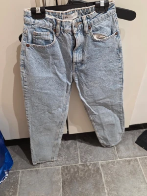 Ljusblå jeans från Zara, storlek 32 - Säljer ett par ljusblå jeans från Zara med klassisk femficksdesign och rå kant upptill. Jeansen har hög midja och raka ben, tillverkade i slitstarkt bomullsjeansmaterial. Snygga kontrastsömmar och metallknapp framtill. Perfekta för en chill och trendig look.