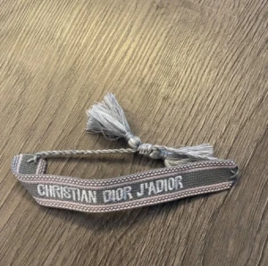Grått Christian Dior J'ADIOR armband - Säljer ett snyggt vävt armband från Christian Dior med texten 'J'ADIOR' i vitt. Armbandet är grått med ljusare detaljer och har tofsar i ändarna. Perfekt accessoar för att lyfta din outfit.