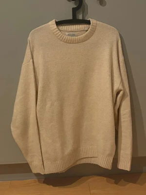 Beige stickad tröja  - Säljer en beige stickad tröja från Jack & Jones med rund hals och ribbade muddar vid ärmslut och nederkant. Tröjan har en avslappnad passform. Tröjan är knappt använd så den är i nyskick.