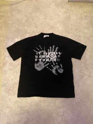 Maison Margiela svart t-shirt med tryck - Maison Margiela svart t-shirt i bomull med unikt vitt tryck av handavtryck och siffror på bröstet. Klassisk passform och rund hals. Tillverkad i Italien och har en clean, edgy vibe som sticker ut.