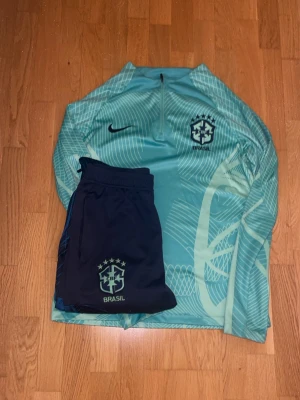 Nike Brasil träningsset blå/grön - Säljer ett Nike Brasil träningsset med långärmad tröja i ljusblått med grafiskt mönster och halv dragkedja samt matchande mörkblå byxor med Brasil-logga och Nike Swoosh. Perfekt för fotbollsträning eller chill. Materialet är syntet och andas bra.
