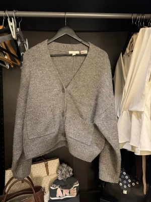Grå stickad kofta från H&M - Mysig grå stickad kofta från H&M med v-ringning och stora knappar framtill. Koftan har två rejäla fickor och en loose passform som ger en chill vibe. Perfekt att slänga på sig över en t-shirt eller topp när det är kyligt ute.
