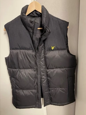 Svart väst från Lyle & Scott - Säljer en svart väst från Lyle & Scott med gul broderad logga på bröstet. Västen har hög krage, quiltad design och stängs med både dragkedja och tryckknappar. Perfekt att slänga på över en hoodie när det är kyligt ute. Bara att skriva för mer info!