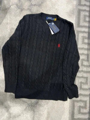 Svart kabelstickad tröja Polo Ralph Lauren - Svart kabelstickad tröja från Polo Ralph Lauren med klassisk rund hals och röd broderad logga på bröstet. Tröjan har ribbade muddar vid ärmslut och nederkant, samt långärmad passform. Perfekt för dig som gillar stilrena och tidlösa plagg. Priset kan självklart diskuteras 😊