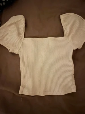 Beige ribbad topp med puffärm H&M XS - Säljer en beige ribbad topp från H&M i storlek XS. Toppen har puffiga korta ärmar och en fyrkantig halsringning. Materialet är mjukt och stretchigt, perfekt för dig som gillar en stilren och trendig look. Passformen är slim och toppen är croppad.