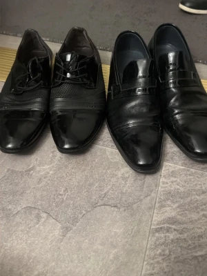 Svarta lackade finskor och loafers150 st - Två par eleganta svarta finskor, ett par med snörning och ett par klassiska loafers. Båda har spetsig tå och blank finish som ger en riktigt dressad look. Perfekta till kostym eller när du vill steppa upp stilen. Materialet är glansigt skinn.