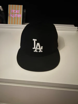 Svart LA New Era 59FIFTY keps - Säljer en klassisk svart LA-keps från New Era, modell 59FIFTY. Kepsen har broderad LA-logga framtill och New Era-logga på sidan, samt MLB-logga bak. Snygg och stilren modell med rak skärm och strukturerad passform. Inte använd