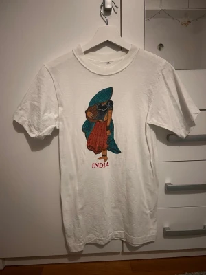 Vit t-shirt med India-tryck - Vit t-shirt i bomull med färgglatt tryck av en indisk kvinna och texten 'INDIA' framtill. Klassisk rund hals och korta ärmar. Perfekt för dig som gillar unika motiv och vill sticka ut lite extra.