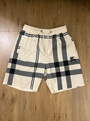 Burberry Badshorts  - Bad byxor/ surfshorts från Burberry. Vit/ljus beige med grå/ svarta detaljer. Inte mycket använda, så är bra! Finns en liten svart fläck på baksidan men inget som syns så mycket. Skickar snabbt