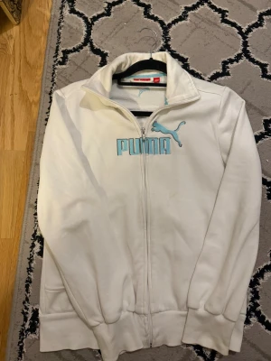 Vit ziptröja från Puma med blå logga - Vit långärmad ziptröja från Puma med ljusblå broderad logga och text på bröstet. Tröjan har ståkrage, två sidofickor och ribbade muddar. Materialet är mjuk bomull med insida i fleece. Perfekt för chill eller sportiga dagar.