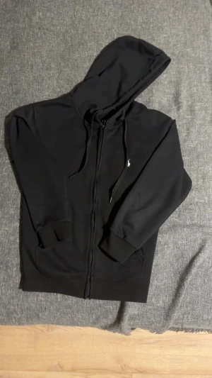 Svart hoodie från Polo Ralph Lauren - Svart zip-hoodie från Polo Ralph Lauren i storlek S. Klassisk design med huva och dragsko, ribbade muddar och fickor framtill. Liten broderad logga på bröstet. Mjuk och skön bomullsblandning som passar perfekt till chill. Köpte den för 2 månader sen men har knappt använt den och nu har den blivit liten på mig. Skicket är helt ny, ni kan se de på bilden, inga fläck, inget smuts och den luktar inget. Original priset är 2000 kr men jag säljer den för 1100 eftersom de inte går att returnera den.