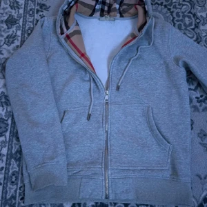Grå Burberry hoodie med dragkedja - Säljer en grå hoodie från Burberry med klassiskt rutigt foder i huvan och broderad logga på bröstet. Tröjan har snörning i huvan, praktiska fickor och hel dragkedja framtill. Perfekt för dig som gillar stilren design med lyxig touch.