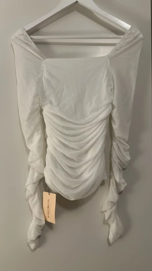 Vit omlott topp med rynk från SHEIN - Supersnygg vit topp från SHEIN med omlott och rynkade detaljer framtill. Långa, puffiga ärmar som ger en cool och trendig vibe. Materialet är lätt och känns skönt mot huden. Perfekt för dig som vill sticka ut med en unik och fräsch look.