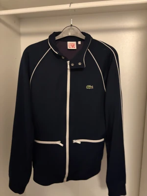 Mörkblå tröja från Lacoste LIVE - Snygg mörkblå tröja från Lacoste LIVE med vita detaljer och dragkedjor. Tröjan har ståkrage med tryckknappar, två fickor med dragkedja och den klassiska Lacoste-krokodilen på bröstet. Perfekt för dig som gillar sportig och clean stil. Ny pris 1200kr