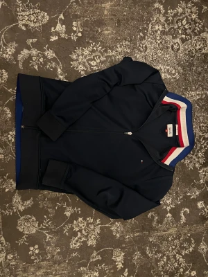 Mörkblå ziptröja Tommy Hilfiger - Snygg mörkblå ziptröja från Tommy Hilfiger med hel dragkedja och ribbade muddar. Kragen har coola ränder i rött, vitt och blått som ger en sportig vibe. Liten Hilfiger-logga på bröstet. Perfekt för dig som gillar stilrena och klassiska plagg.