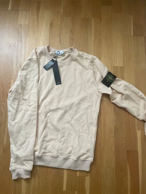 Beige sweatshirt från Stone Island - Snygg beige sweatshirt från Stone Island med klassisk rund hals och ribbade muddar. Tröjan har den ikoniska Stone Island-loggan på vänster ärm och är tillverkad i mjuk bomull. Perfekt för dig som gillar stilrena och cleana plagg med streetkänsla.