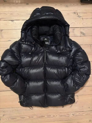 Svart dunjacka från J.Lindeberg - Säljer en svart puffer från J.Lindeberg med stor huva och markerad logga på ärmen. Den är rejält vadderad med 100% dun (Duck feather) som ger otroligt bra värme. Glansig finish som ger en stilig look🙌🏻 Köpt förra året, sparsamt använd och i helt perfekt skick! 