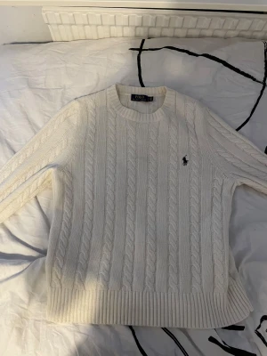 Vit kabelstickad tröja Polo Ralph Lauren - Vit kabelstickad tröja från Polo Ralph Lauren med klassisk rund halsringning och broderad logga på bröstet. Tröjan är i mjuk bomull och har ribbade muddar vid ärmslut och nederkant. Perfekt för en clean och stilren look.