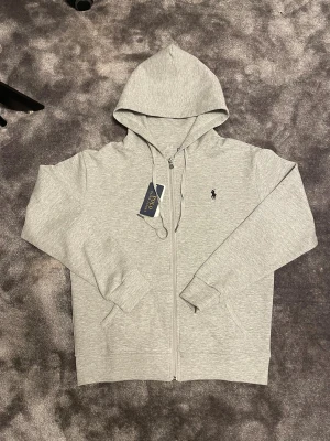 Grå hoodie zip up - Snygg ljusgrå hoodie  med dragkedja och huva med snörning. Tröjan har långärmade ribbade muddar och klassisk Polo-logga broderad på bröstet. Perfekt för chill dagar och enkel att matcha med jeans eller joggers. Har en i M också