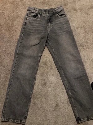Grå raka jeansbyxor - Säljer ett par grå jeansbyxor med rak passform och klassisk femficksdesign. Byxorna har en mörkgrå tvättad look och är tillverkade i slitstarkt denim. Perfekt för dig som gillar en enkel och stilren vibe.