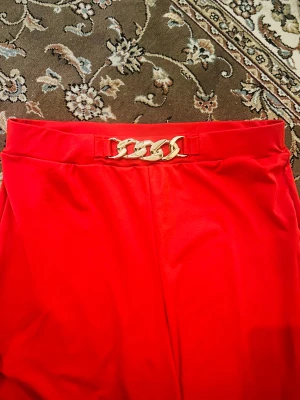 Röda wide pants från SHEIN med kedjedetalj - Snygga röda byxor från SHEIN i storlek 0XL, tillverkade i mjuk och stretchig polyester med 5% elastan. Modellen är wide leg och har en hög midja med en cool guldfärgad kedjedetalj framtill. Perfekt för dig som gillar att sticka ut med färg.