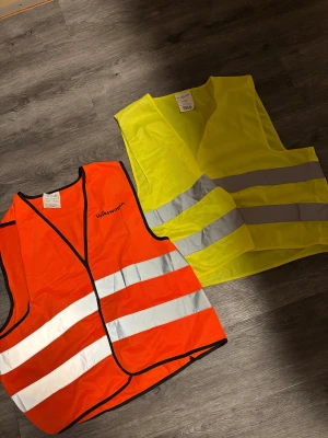 Reflexvästar Volkswagen & RFX Care  - Två reflexvästar, en orange med Volkswagen-tryck och en gul från RFX Care. Båda har breda reflekterande ränder runt midjan och är tillverkade i 100% polyester. Perfekt när du vill synas extra mycket ute eller på jobbet. 