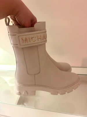 Beige boots från Michael Kors - Säljer ett par snygga boots från Michael Kors i beige skinn med grov sula och guldfärgade detaljer. Stövlarna har ett brett band med MICHAEL i guld framtill och MK-logga bak. Stilren design med rund tå och platt sula, perfekta för att lyfta din look.