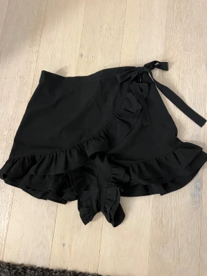 Svarta omlottshorts med volang - Säljer ett par svarta omlottshorts med volangkant och knytband i midjan. De har en feminin och trendig look med volanger längs nederkanten och omlott-detalj framtill. Perfekta för dig som vill ha något snyggt och bekvämt till sommaren.