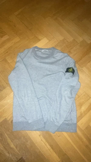 Grå Stone Island sweatshirt M - Säljer en grå sweatshirt från Stone Island i storlek M. Tröjan har klassisk rund hals, ribbade muddar och den ikoniska Stone Island-loggan på vänster ärm. Tillverkad i mjuk bomull för skön passform och stilren look.