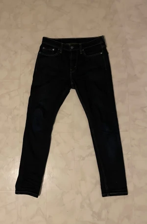 Levi's mörblåa jeans W29 L30 - Snygga mörkblåa Levi's jeans med klassisk femficksdesign och gul kontrastsöm. Modellen är smal och sitter tight längs benen, perfekt för dig som gillar en modern och stilren look. Jeansmaterialet är slitstarkt och har en cool finish.