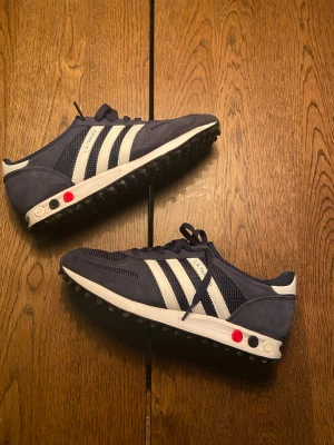 Adidas LA Trainer blå sneakers - Säljer ett par Adidas LA Trainer sneakers i marinblått med vita ränder och detaljer. Skorna har snörning, platt sula med gummidubbar och är tillverkade i mocka och syntet. Klassisk retrostil med coola färgdetaljer på sulan. Perfekt för dig som gillar sportig och tidlös look.