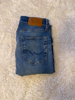 Blå jeans från Jack & Jones - Snygga blå jeans från Jack & Jones med klassisk femficksdesign och läderpatch bak i midjan. Jeansen är i slim fit och subtila slitningar framtill och baktill för en cool, avslappnad look. Perfekt för dig som gillar stilrena och tidlösa jeans.