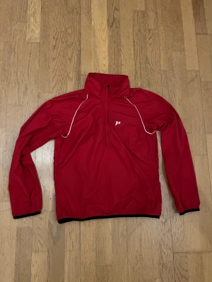 Röd löparjacka från Peak Performance - Peak Performance vindjacka i rött med vita detaljer längs axlarna och halv dragkedja framtill. Jackan har hög krage, elastiska muddar och diskret logga på bröstet. Materialet är lätt och syntetiskt, perfekt för utomhusaktiviteter. Snygg och sportig look.