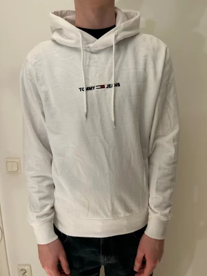 Vit hoodie från Tommy Jeans - Snygg vit hoodie från Tommy Jeans med broderad logga på bröstet. Tröjan har huva med snörning, ribbade muddar och är tillverkad i mjuk bomull. Perfekt för chill dagar och enkel att matcha med jeans eller joggers.