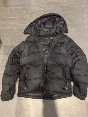 Svart pufferjacka Polo Ralph Lauren - Svart pufferjacka från Polo Ralph Lauren med huva och två sidofickor med dragkedja. Jackan har en röd Polo-logga på bröstet och quiltad design. Inga hål. Nypris är 5995, QR kod och kvitto finns! Priset är fast.