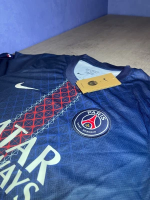 PSG x Nike blå fotbollströja - Säljer en officiell Paris Saint-Germain t-shirt från Nike i marinblått med geometriskt mönster i rött och vitt längs mitten. PSG-logga och Nike Swoosh på bröstet, samt Qatar Airways-tryck. Tröjan är kortärmad och gjord i lätt polyester, perfekt för fotboll.
