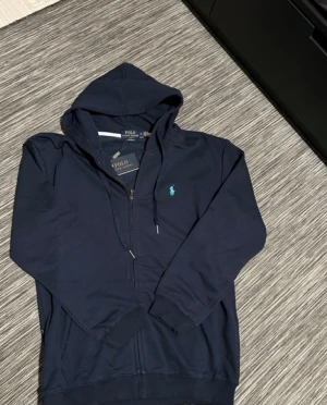Mörkblå hoodie från Polo Ralph Lauren - Snygg mörkblå hoodie från Polo Ralph Lauren med dragkedja och huva. Klassisk logga i ljusblått på bröstet, ribbade muddar och fickor framtill. Tillverkad i mjuk bomull för skön komfort. Perfekt för chill dagar och streetwear-stil.