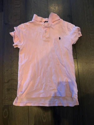 Rosa pikétopp från Polo Ralph Lauren - Säljer en ljusrosa pikétopp från Polo Ralph Lauren med klassisk krage, korta ärmar och knappar framtill. Toppens material är bomull och har den ikoniska broderade loggan i svart på bröstet. Perfekt för en chill och sportig look.