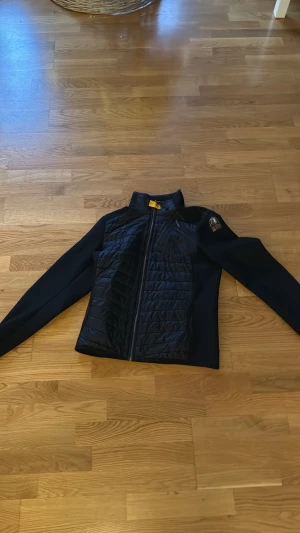 Svart cardigan jacka från Parajumpers - Svart jacka från Parajumpers med quiltad front och rygg, samt släta ärmar. Jackan har dragkedja fram och en snygg patch på ena ärmen. Materialmix med polyester och syntet, perfekt för kyliga dagar. Snygg och sportig look med coola detaljer. Dm för fler bilder och kan sänka pris men inte för mycket.