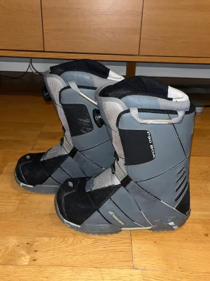 Grå snowboardboots BOA 26cm - Säljer ett par grå och svarta snowboardboots från LH med BOA-snörning. Bootsens ovandel är i syntetmaterial med robust sula och förstärkta partier. De har dragöglor upptill för enkel påtagning och en sportig look som passar vinterns åk. 26cm lång